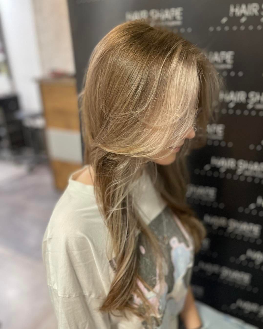 Długie włosy z jasnym balejażem na tle ściany z logo Hair Shape